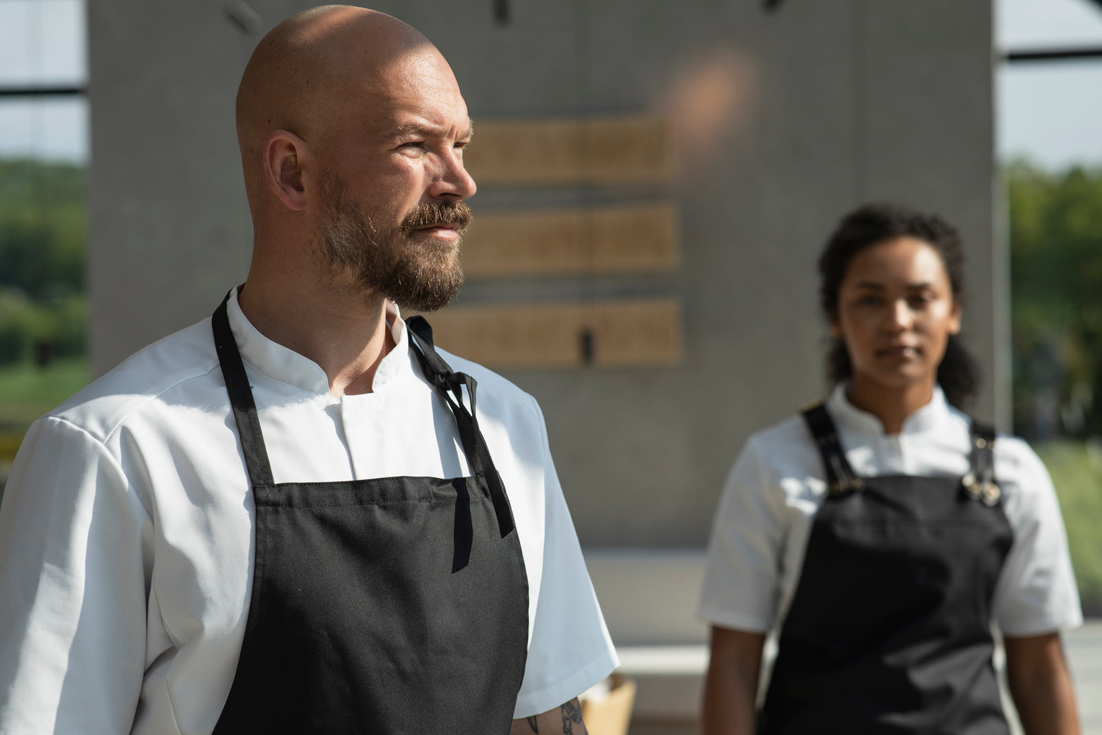 Två professionella kockar i Segers bröstlappsförkläden, manlig kock i förgrunden med ett klassiskt svart förkläde och kvinnlig kock i bakgrunden med ett svart förkläde med hängslen, arbetar i ett ljust och modernt kök.